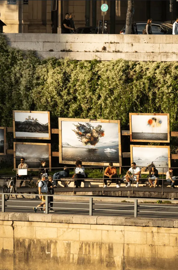 Des photographies grand format sont exposées en plein air le long d’un quai, au-dessus d’une rangée de bancs où des passants sont assis ou se promènent. Justine Brunet