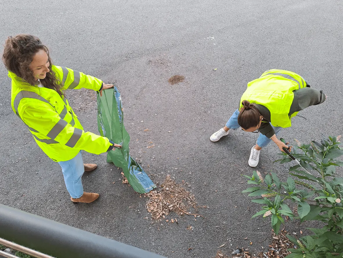 Deux personnes en gilets fluorescents ramassent des déchets au sol, l’une tenant un sac ouvert tandis que l’autre utilise une pince pour collecter les détritus.