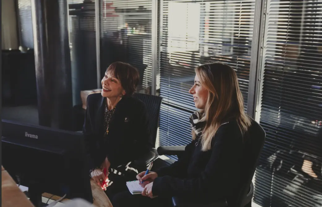 Deux femmes sont assises côte à côte dans un bureau, regardant un écran et prenant des notes dans une ambiance de travail collaborative. Alexandre Bagdassarian