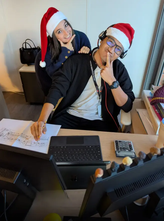 Deux collègues portent des bonnets de Noël et posent avec humour à leur poste de travail, entourés d’écrans et de documents.
