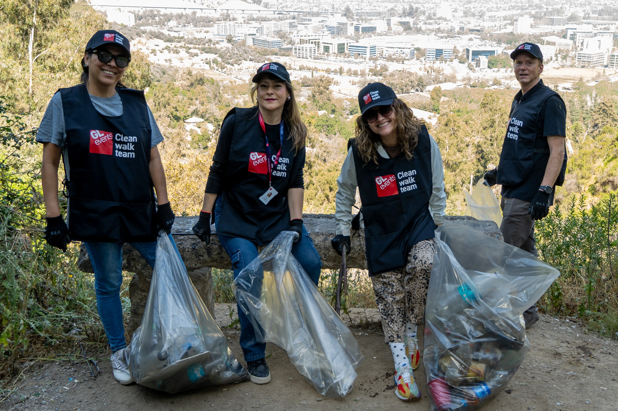 Quatre bénévoles portant des gilets « GL events Clean Walk Team » posent en plein air avec des sacs remplis de déchets ramassés, surplombant un paysage urbain.