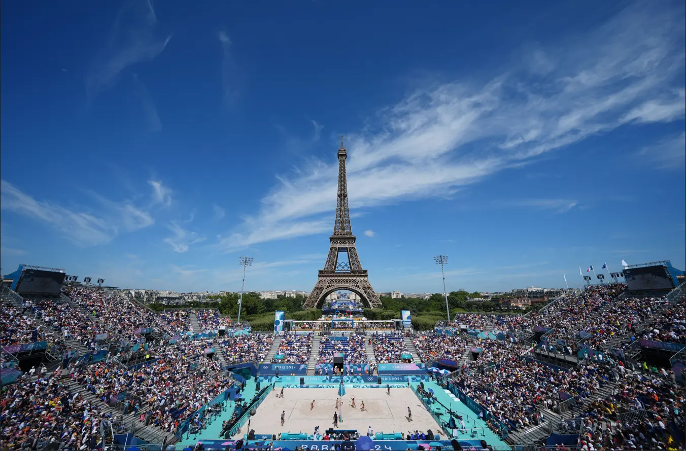 Stade temporaire de beach-volley bondé installé au pied de la tour Eiffel à Paris, avec des tribunes pleines de spectateurs sous un ciel bleu dégagé.