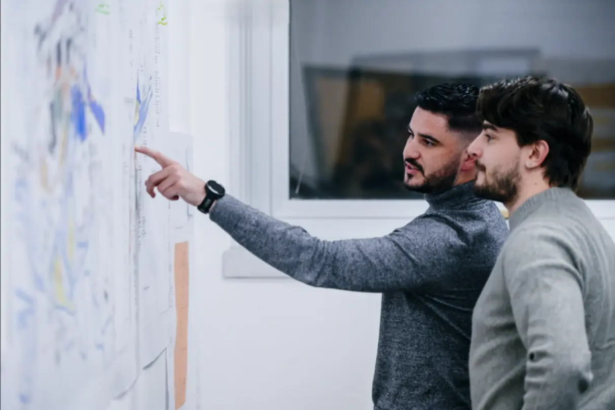 Deux collaborateurs analysant des plans affichés au mur, l’un pointant un détail tandis qu’ils échangent dans un environnement de bureau. Mahdi ARIDJ