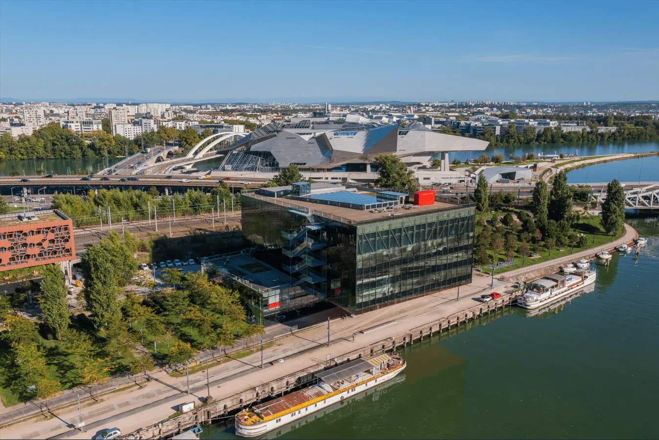Vue aérienne d’un quartier urbain en bord de rivière, avec un bâtiment moderne en verre au premier plan, des péniches amarrées et, en arrière-plan, un grand complexe architectural contemporain relié par un pont. TrenteSept 