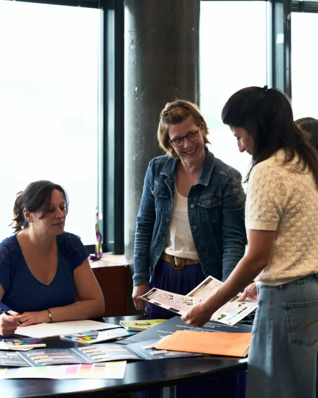 Trois collaboratrices échangeant autour de documents et d’échantillons sur une table, dans un espace de travail lumineux.