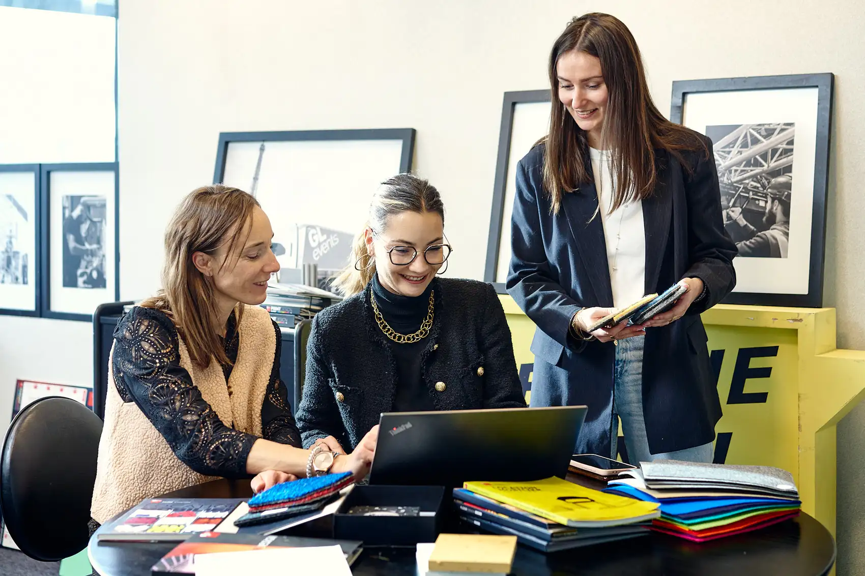 Trois collaboratrices échangeant autour d’un ordinateur portable et d’échantillons de matériaux, dans un environnement de travail créatif.