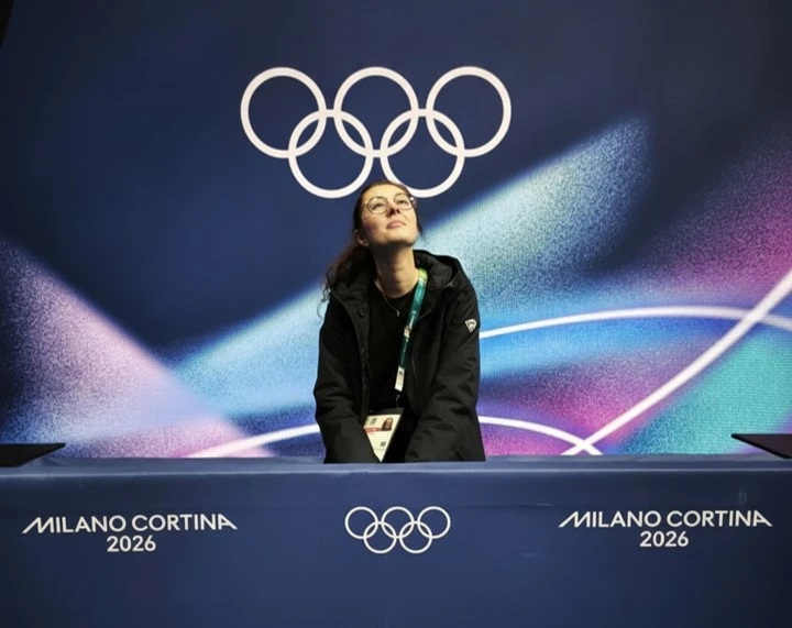 Une femme accréditée, debout derrière un pupitre, regarde vers le haut devant un fond aux couleurs dégradées avec les anneaux olympiques. Le pupitre affiche “Milano Cortina 2026”, faisant référence aux Jeux olympiques d’hiver.