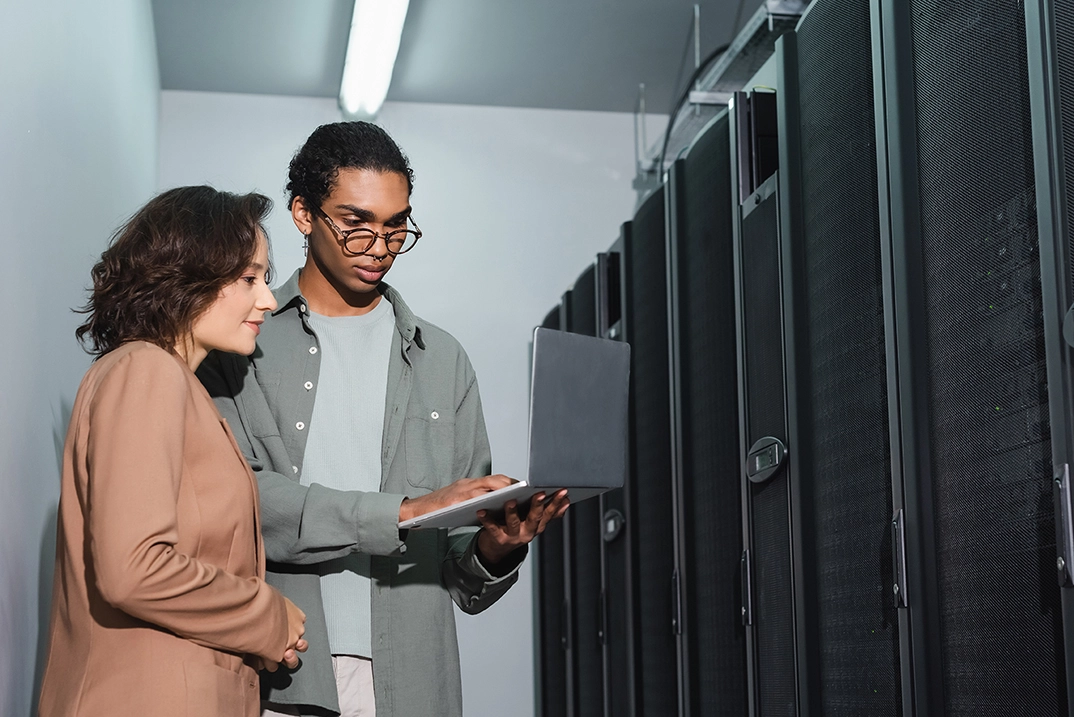 Deux collègues travaillent ensemble dans une salle serveur, consultant un ordinateur portable devant des rangées d’équipements informatiques.