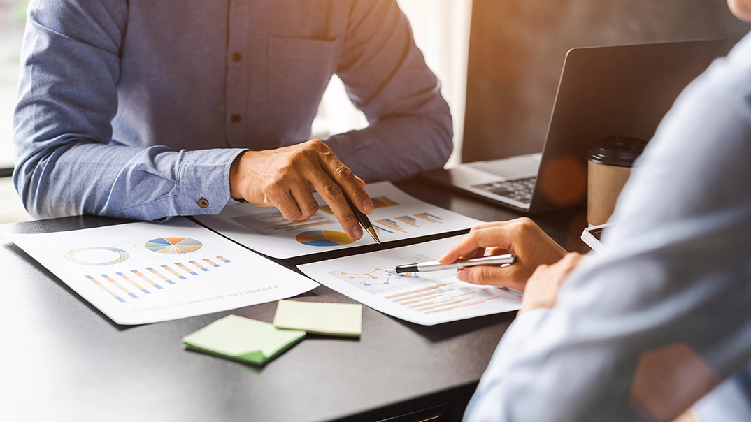 Deux personnes analysent ensemble des graphiques et des documents financiers posés sur une table, en discutant autour d’un ordinateur portable.