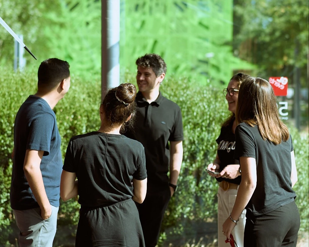 Un groupe de personnes échange dans un espace vitré lumineux, entouré de verdure visible à l’extérieur, dans une ambiance informelle et collaborative.