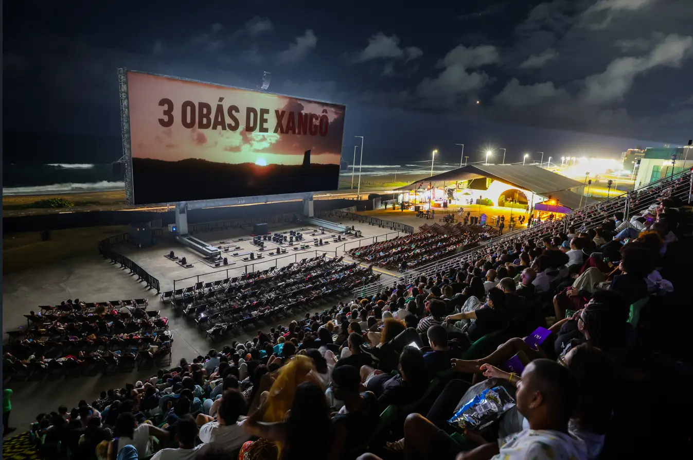 Un cinéma en plein air en bord de mer accueille un large public assis dans des gradins, face à un écran géant diffusant un film de nuit. Fernando Souza
