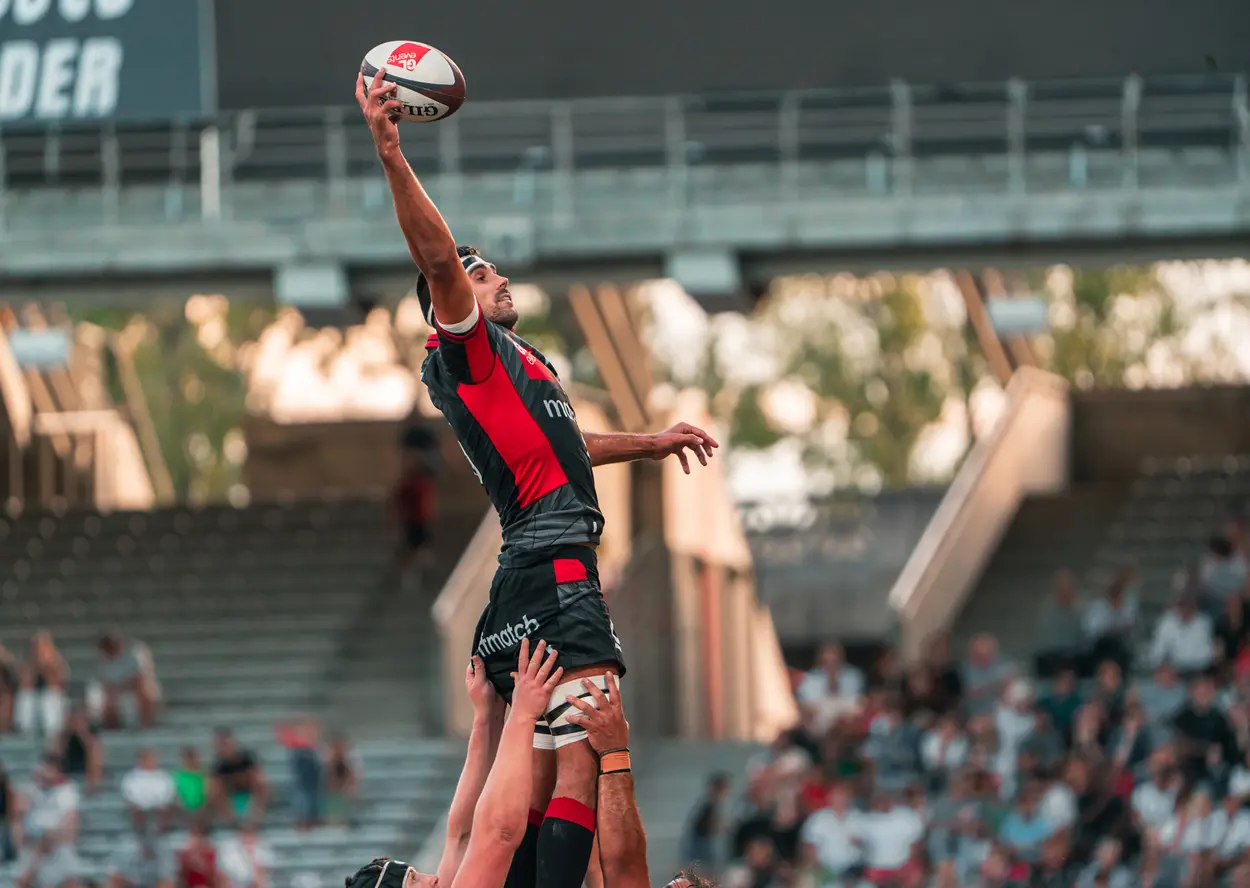 Un joueur de rugby est soulevé par ses coéquipiers lors d’une touche pour attraper le ballon, devant des tribunes remplies de spectateurs.
