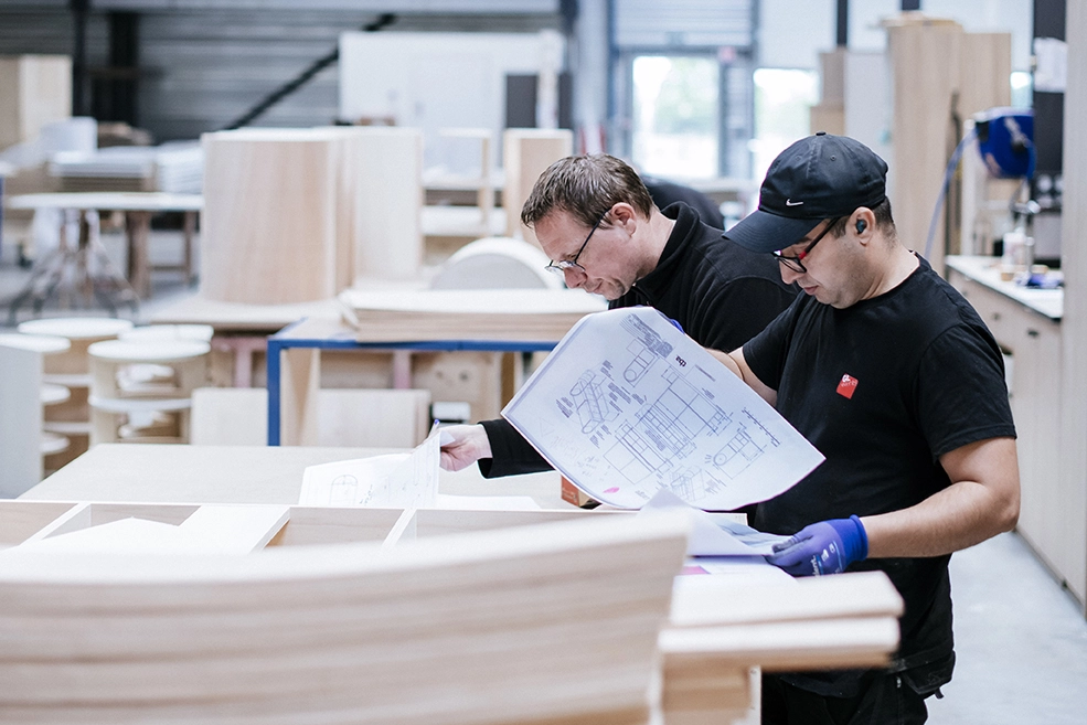 Deux techniciens consultent des plans dans un atelier de menuiserie, entourés de pièces en bois en cours de fabrication.