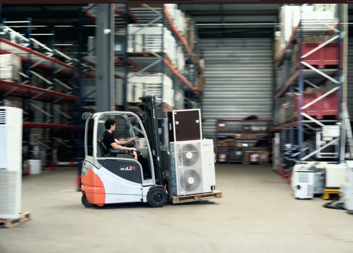 Cariste conduisant un chariot élévateur dans un entrepôt, transportant une palette de matériel entre des rayonnages industriels remplis de marchandises.