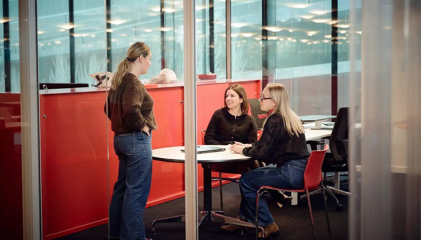 Trois collaboratrices échangent dans un espace de réunion vitré, dans une ambiance détendue et professionnelle. L’une est debout tandis que les deux autres sont assises autour d’une table, illustrant un moment de discussion informelle et de collaboration au quotidien.