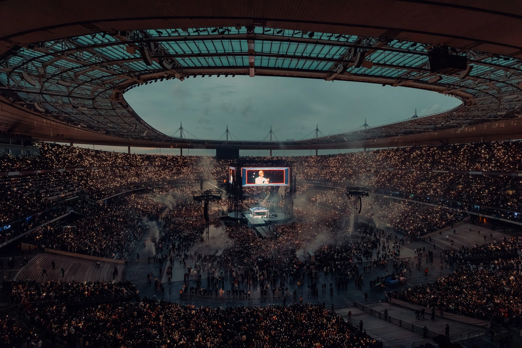 Vue spectaculaire d’un stade rempli lors d’un concert ou événement live, baigné de lumières rouges, avec une foule dense et des milliers de points lumineux créant une ambiance immersive et festive. Nicko Guihal