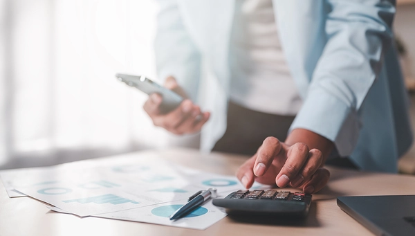 Une personne utilise une calculatrice tout en consultant des documents et un téléphone, dans un contexte d’analyse ou de gestion financière.