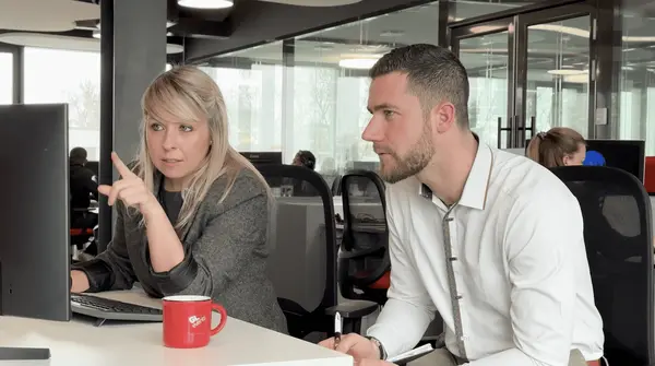 Deux collaborateurs sont concentrés devant un écran dans un open space moderne. La femme, en train de montrer quelque chose du doigt, semble expliquer ou guider, tandis que son collègue écoute attentivement avec un carnet en main. La scène illustre un moment de travail collaboratif, d’analyse et de prise de décision. GL events