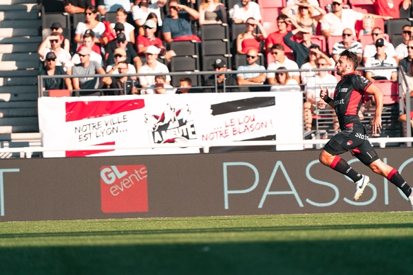 Un joueur de rugby en tenue noire et rouge court sur le terrain pendant un match, devant des tribunes remplies de spectateurs. Des banderoles de supporters et un panneau “GL events” sont visibles en arrière-plan.