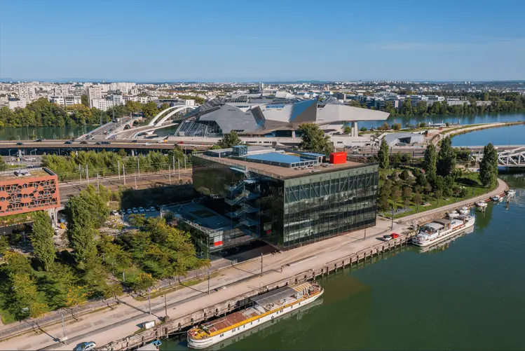 Vue aérienne d’un quartier urbain en bord de rivière, avec un bâtiment moderne en verre au premier plan, des péniches amarrées et, en arrière-plan, un grand complexe architectural contemporain relié par un pont. TrenteSept 