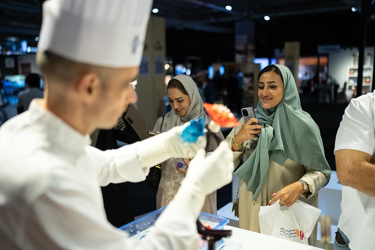 Un chef réalise une préparation culinaire sous les yeux de visiteurs qui observent et prennent des photos lors d’un salon.