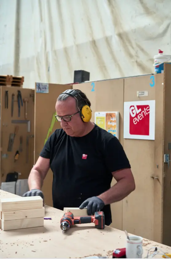 Technicien équipé de protections auditives travaillant le bois sur un établi, dans un atelier, avec des outils et un logo GL events en arrière-plan.