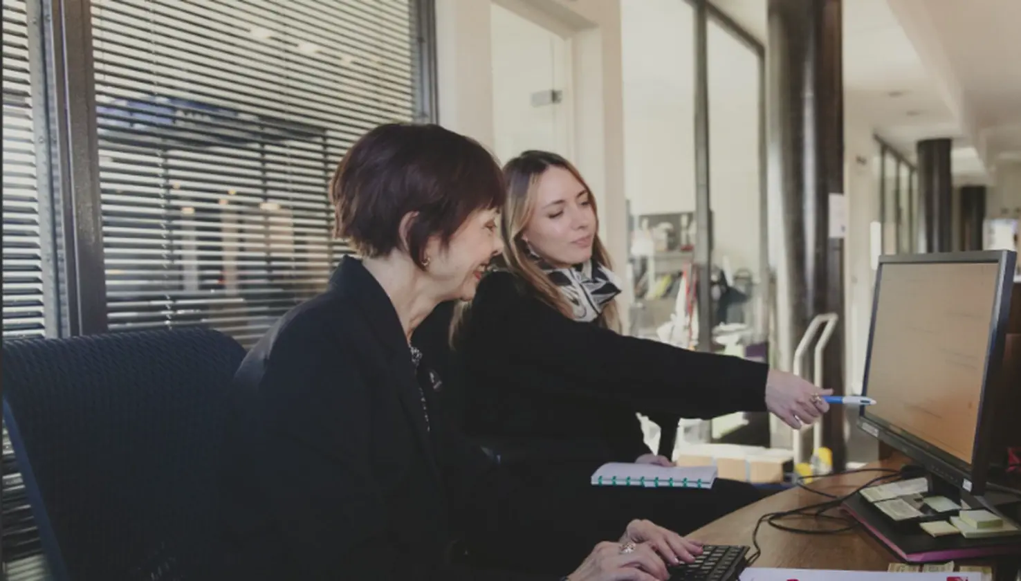 Deux femmes travaillent côte à côte à un bureau d’accueil, l’une utilisant un clavier tandis que l’autre lui montre des informations à l’écran. Alexandre Bagdassarian