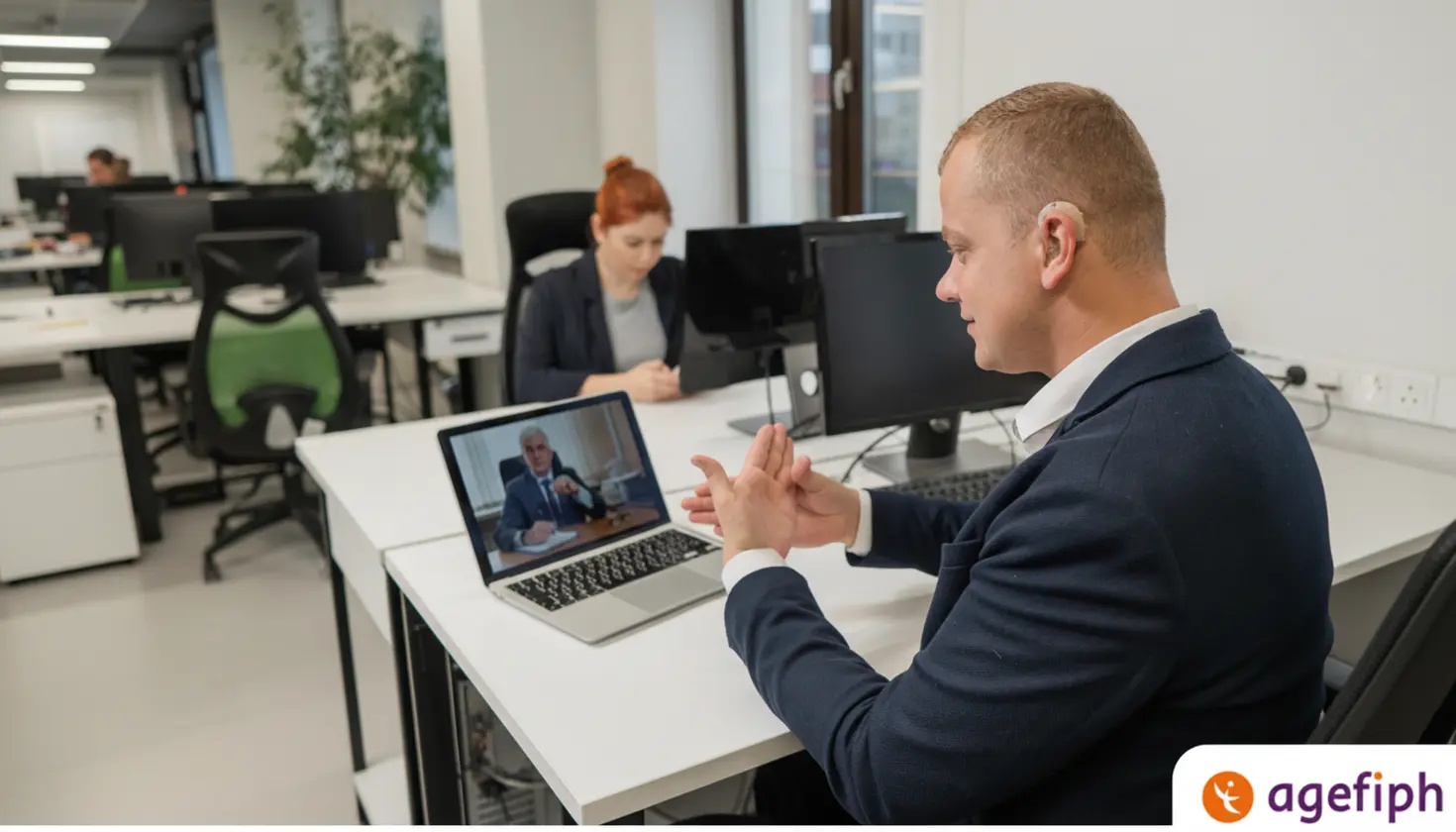 Un homme équipé d’un appareil auditif participe à une visioconférence sur son ordinateur portable dans un bureau, tandis que des collègues travaillent en arrière-plan.