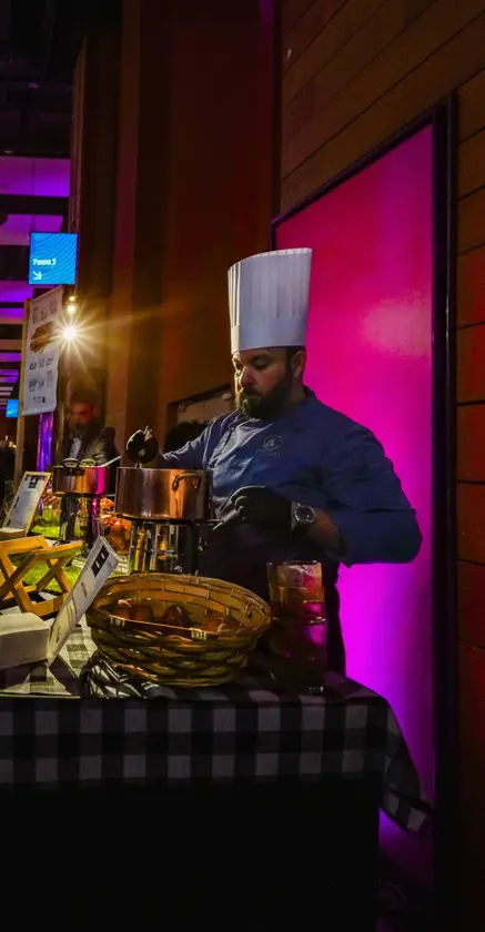 Un chef cuisinier en toque prépare un plat dans une casserole lors d’un événement, devant un stand culinaire éclairé de lumières colorées, avec des ingrédients et des ustensiles disposés sur le plan de travail.