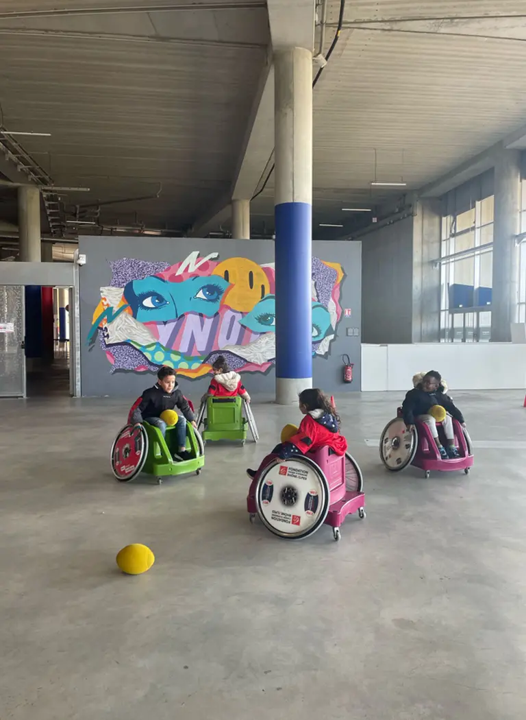 Des enfants jouent ensemble dans un espace intérieur, assis dans des fauteuils roulants colorés, avec des ballons jaunes au sol et une fresque murale colorée en arrière-plan.