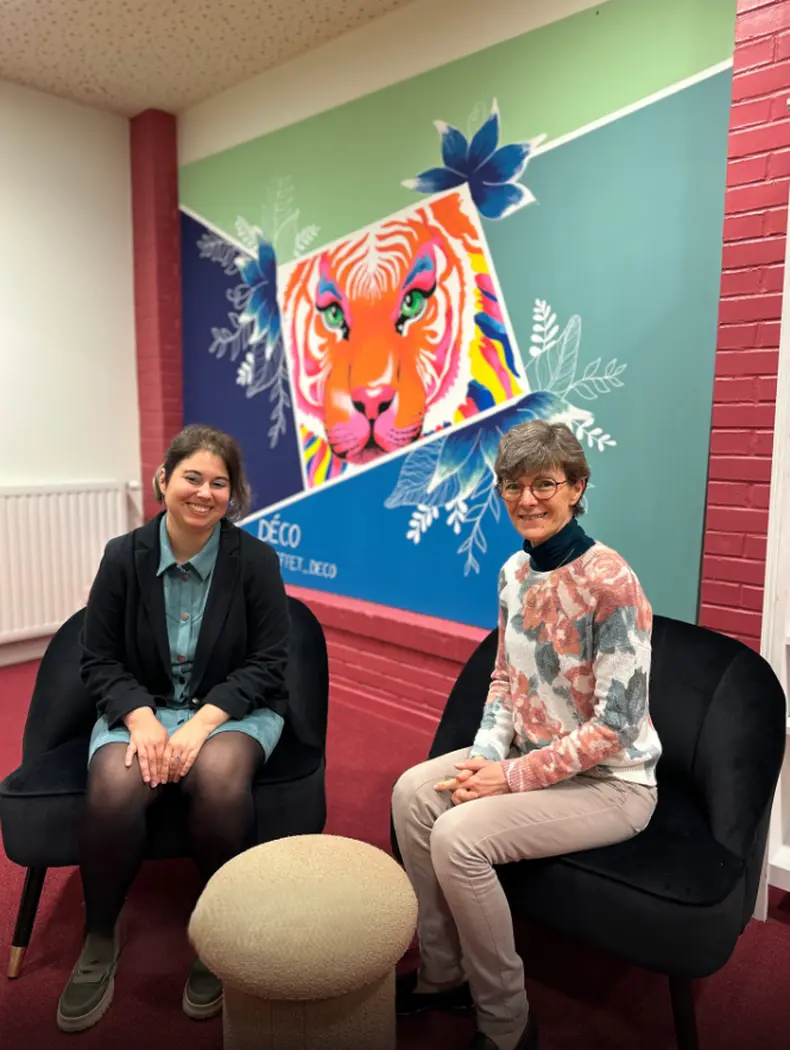 Deux femmes sont assises dans un espace intérieur coloré, devant une fresque murale représentant un tigre stylisé, dans une ambiance conviviale.