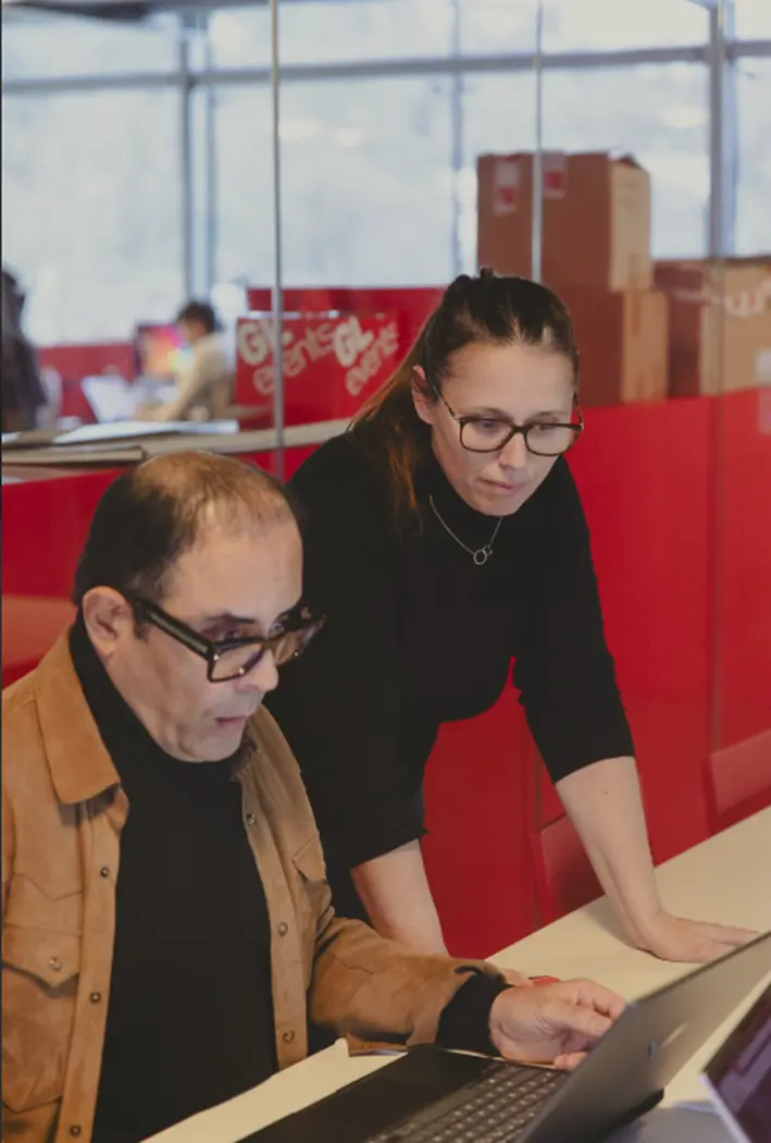 Deux collègues travaillent ensemble autour d’une table, chacun avec un ordinateur portable, dans un espace de bureau moderne aux parois vitrées rouges. Alexandre Bagdassarian