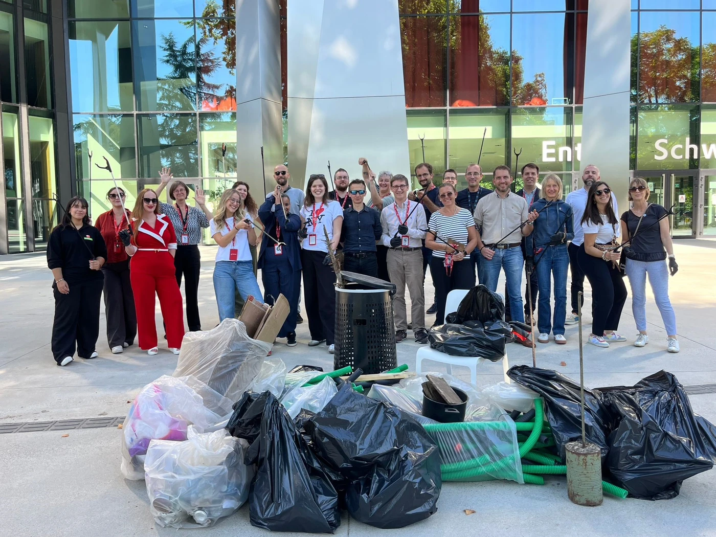 Un groupe de personnes pose devant un bâtiment vitré après une opération de nettoyage, entouré de sacs de déchets et d’outils de ramassage.