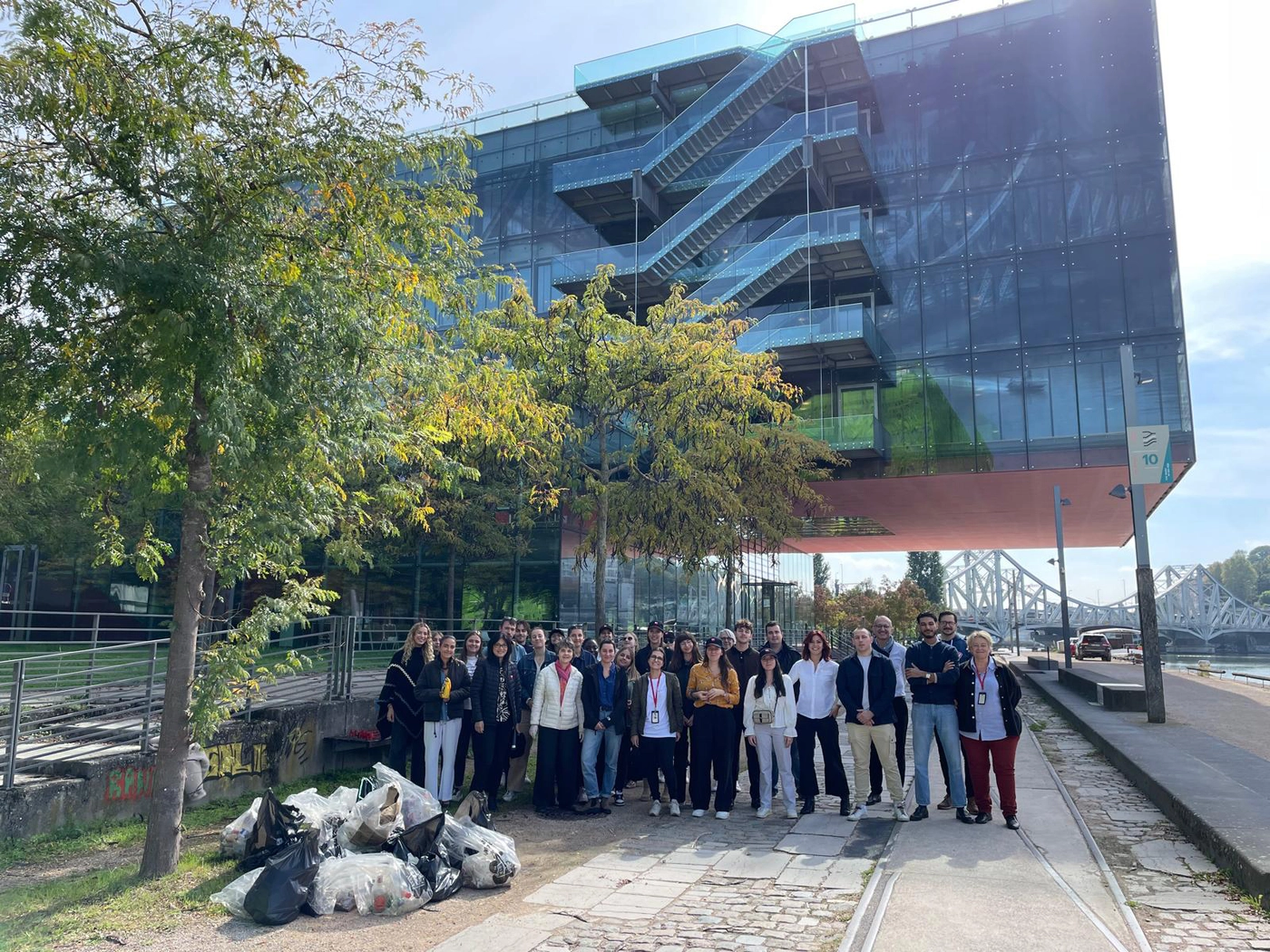 Un groupe de personnes pose au bord d’un fleuve devant un bâtiment moderne, avec des sacs de déchets collectés au premier plan.