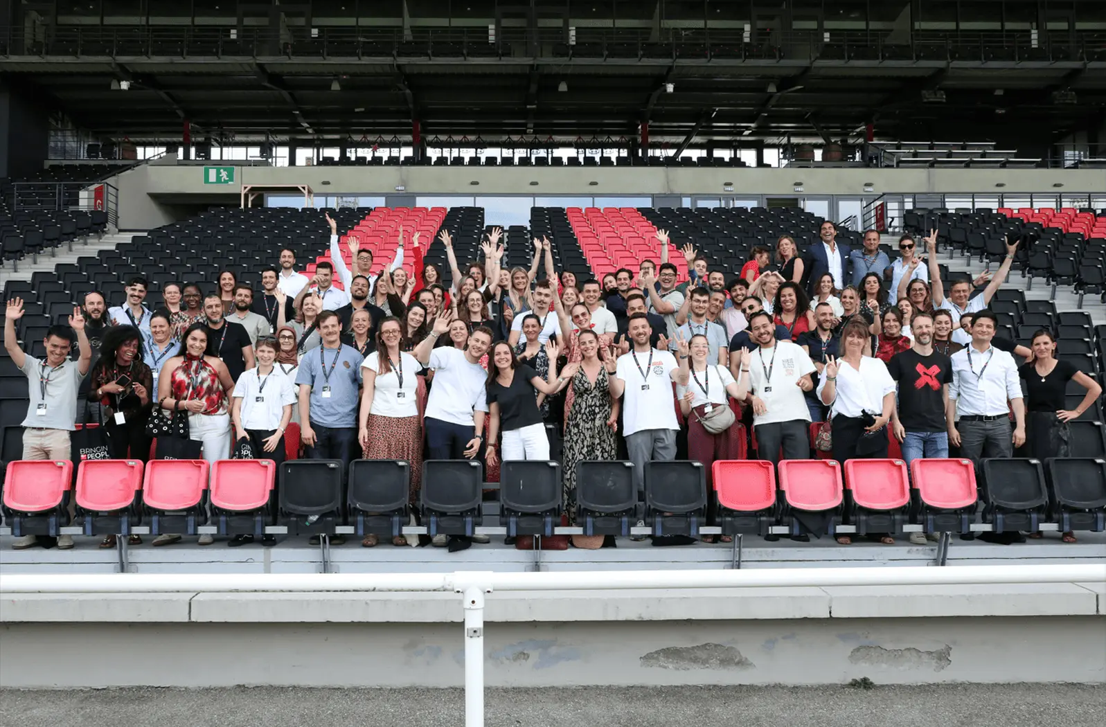 Un grand groupe de collaborateurs pose dans les tribunes d’un stade, souriant et levant les bras dans une ambiance conviviale, illustrant un moment de rassemblement et d’esprit d’équipe. Barbara Tournaire