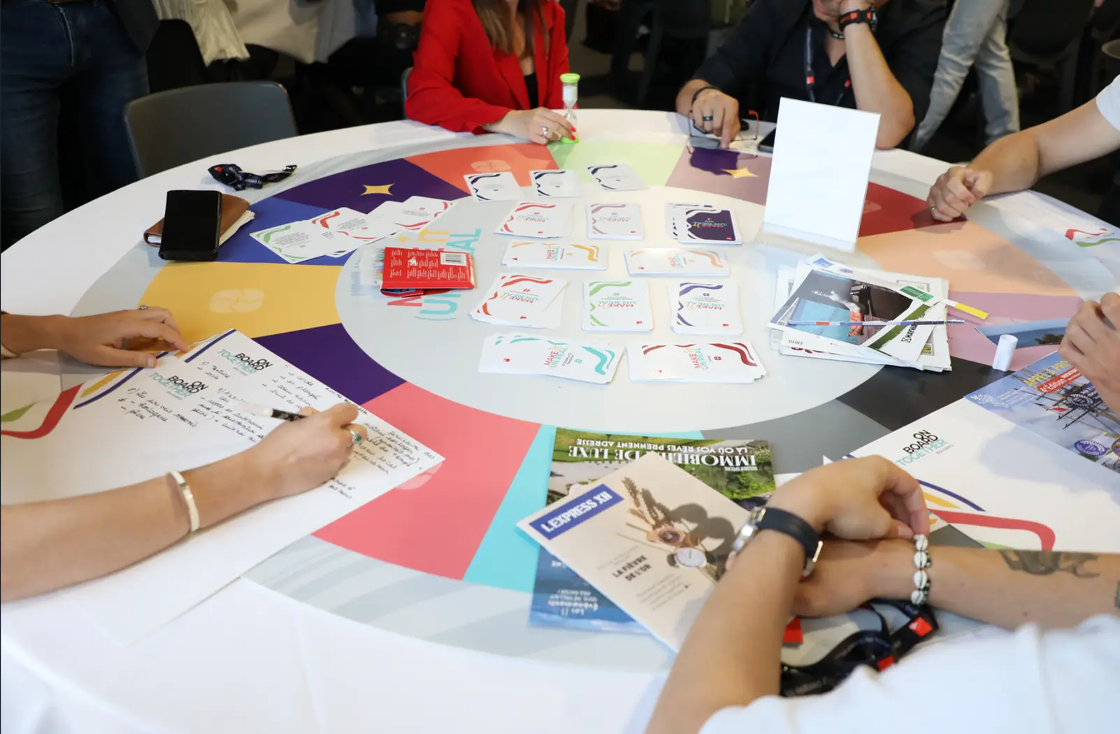 Des collaborateurs participent à un atelier collaboratif autour d’une table ronde, utilisant des cartes, des documents et des supports de travail pour échanger des idées dans le cadre d’une activité de groupe. Barbara Tournaire