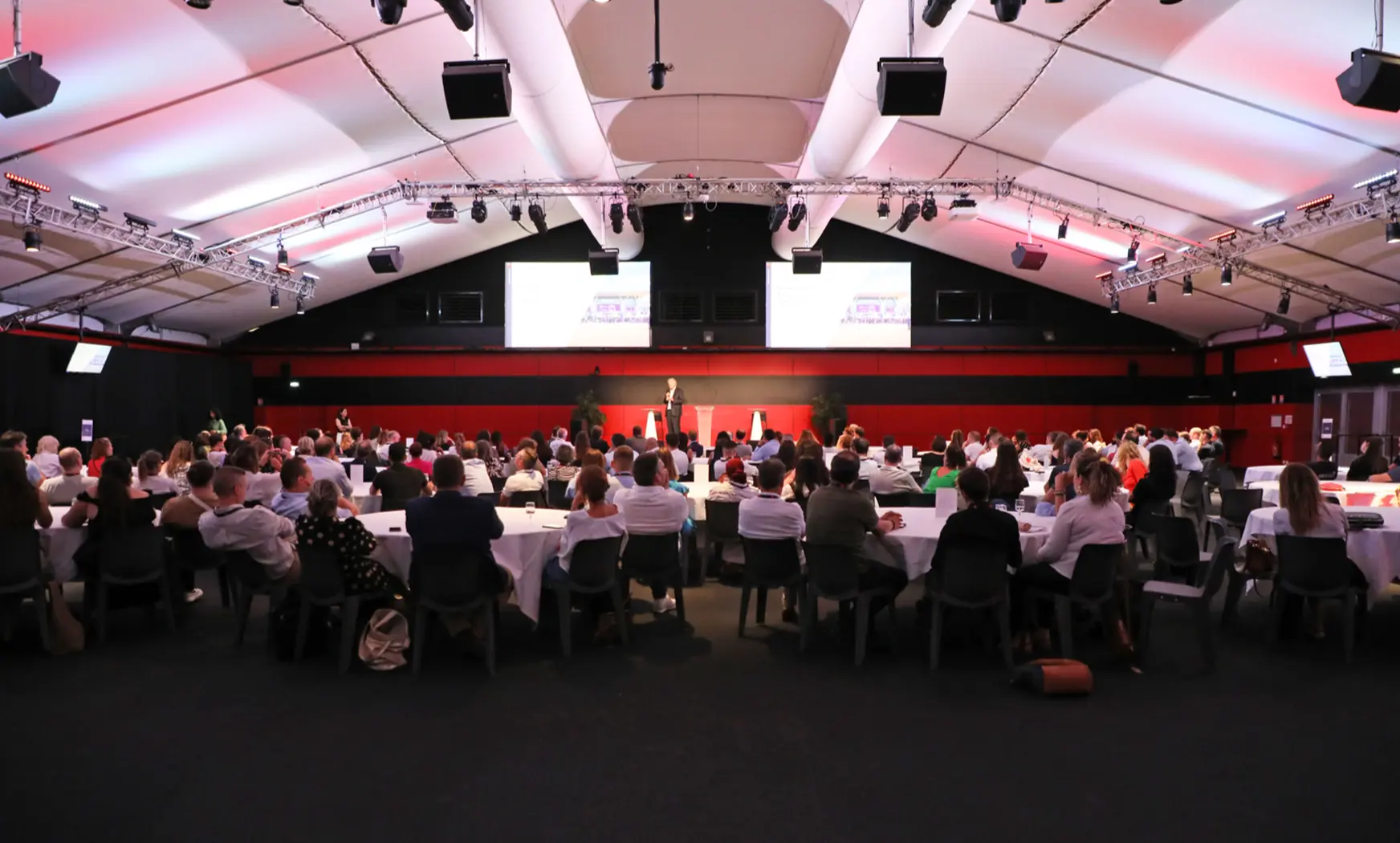Des collaborateurs sont réunis autour de tables rondes dans une grande salle de conférence, assistant à une présentation sur scène diffusée sur deux écrans, lors d’un événement interne ou d’une réunion d’entreprise. Barbara Tournaire